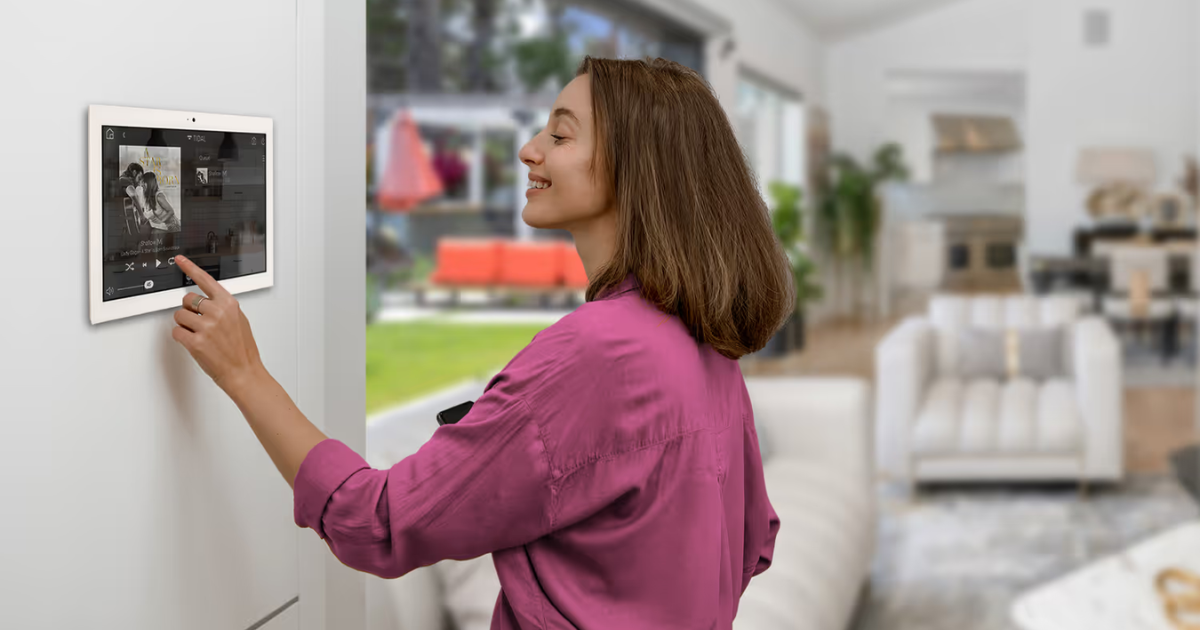 "Woman in a pink shirt smiling while using a wall-mounted smart home touchscreen panel to control music in a bright modern living room.